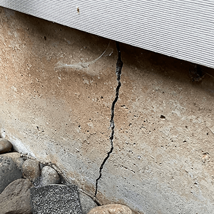 Vertical crack running through a concrete foundation wall with river rock and siding visible above