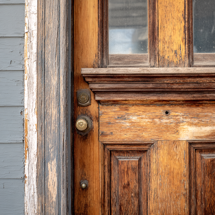 Weathered wooden panel door with original brass hardware and peeling paint on the surrounding frame
